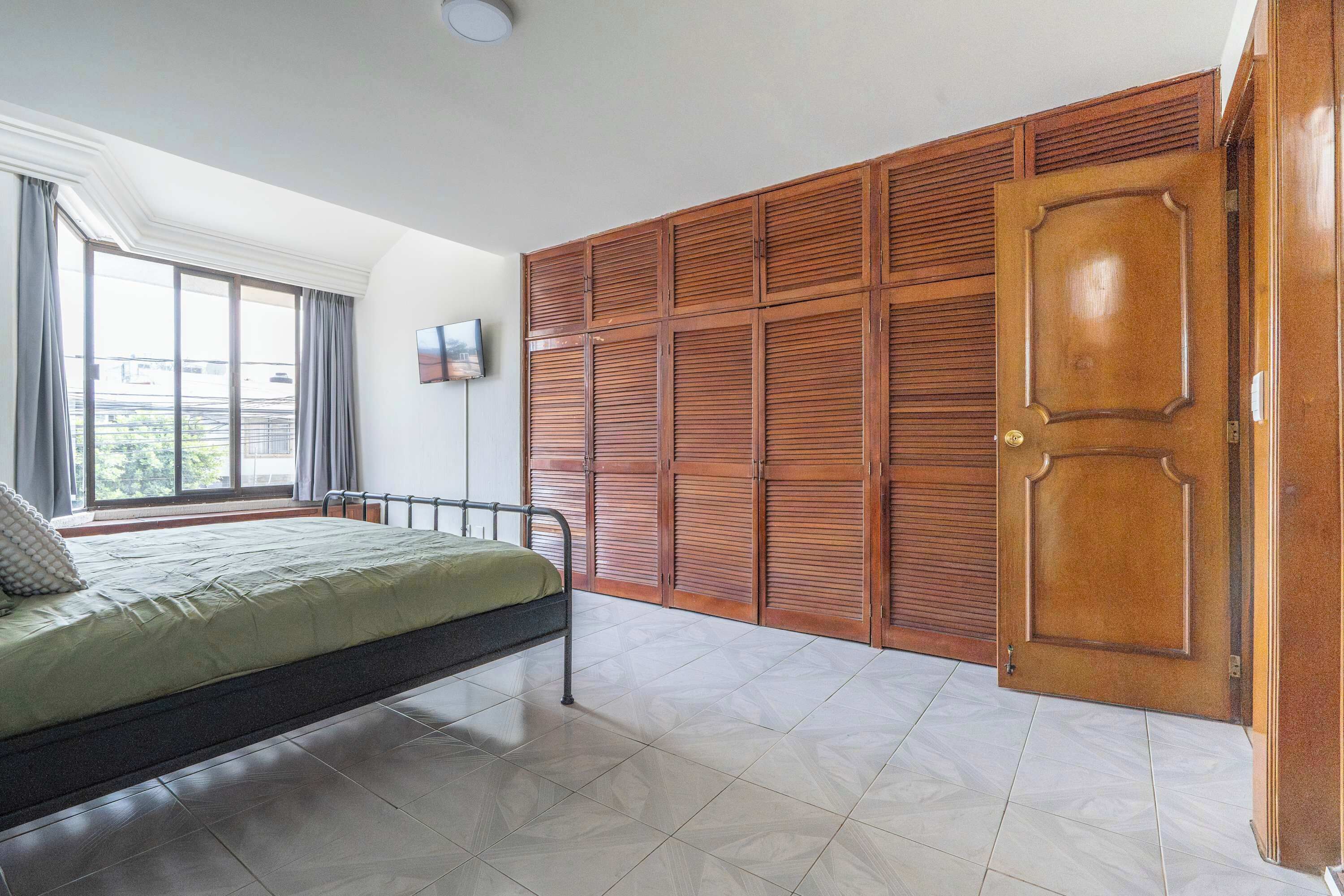 Un dormitorio con cama, armario de madera, ventana con cortinas y televisor de pantalla plana en la pared.