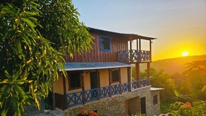 Un bâtiment en bois à deux étages, doté d’un balcon, entouré d’une végétation luxuriante et bordé d’un sentier en pierre.
