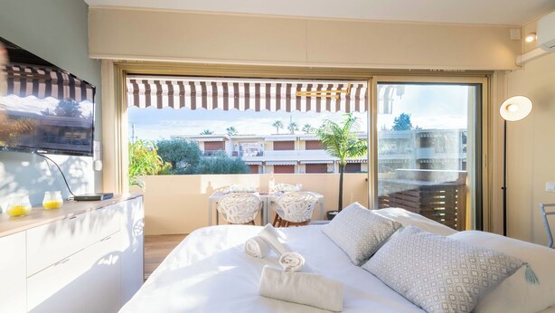Habitación de hotel con una cama grande, televisión, balcón con vistas a edificios y árboles, y una mesa con sillas.