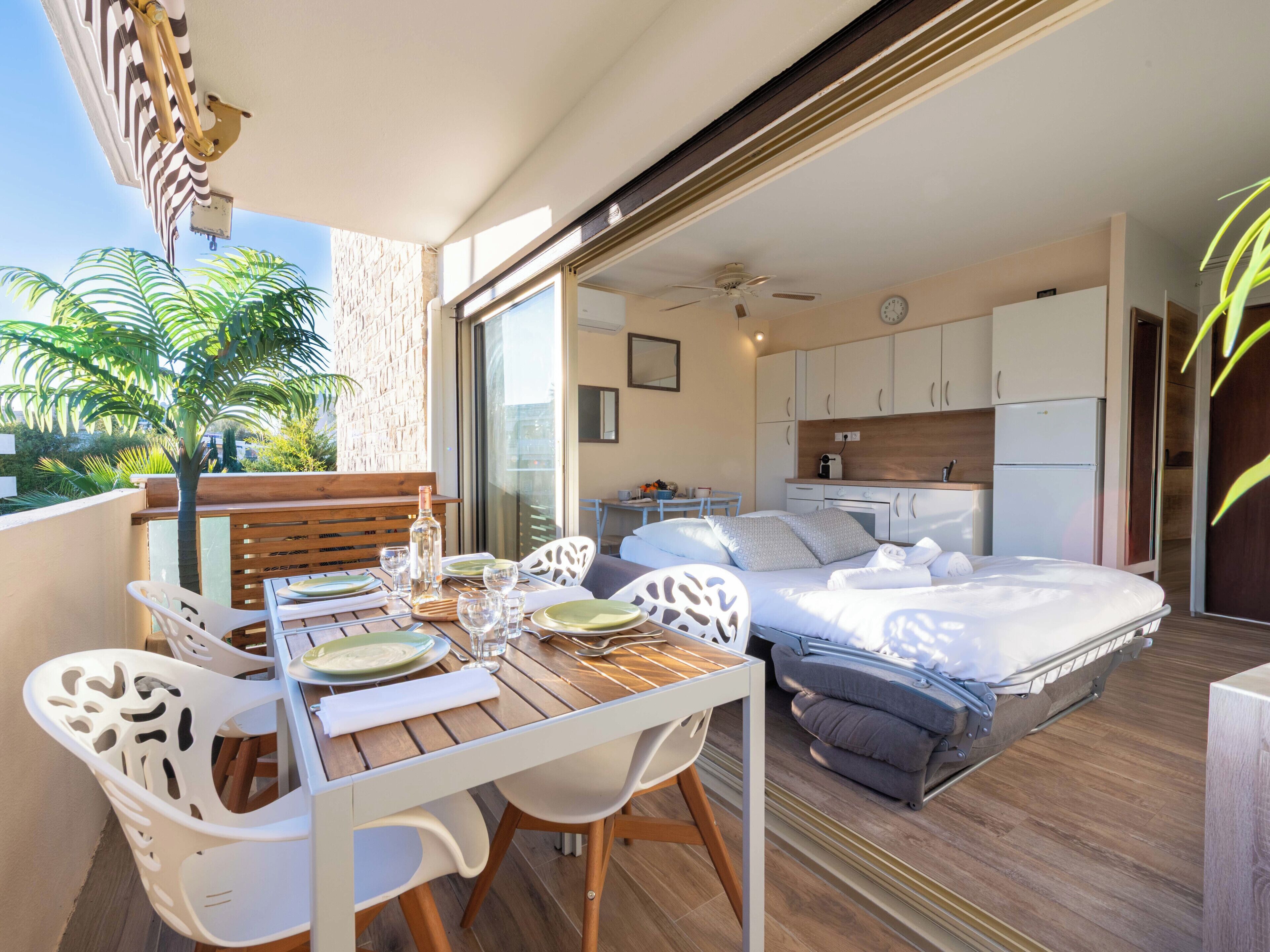 A balcony with a dining table set for two, a bed with a grey mattress, and a kitchen area with white cabinets.