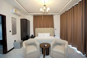 A hotel room with a large bed, two armchairs, a small table, a chandelier, curtains, an air conditioning unit, and a painting on the wall. - Newport Hotel (Atimpoku)