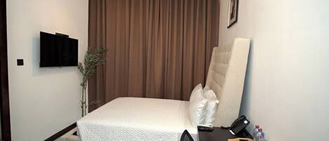 A hotel room with a bed, a desk, a chair, a telephone, a plant, and a TV on the wall.