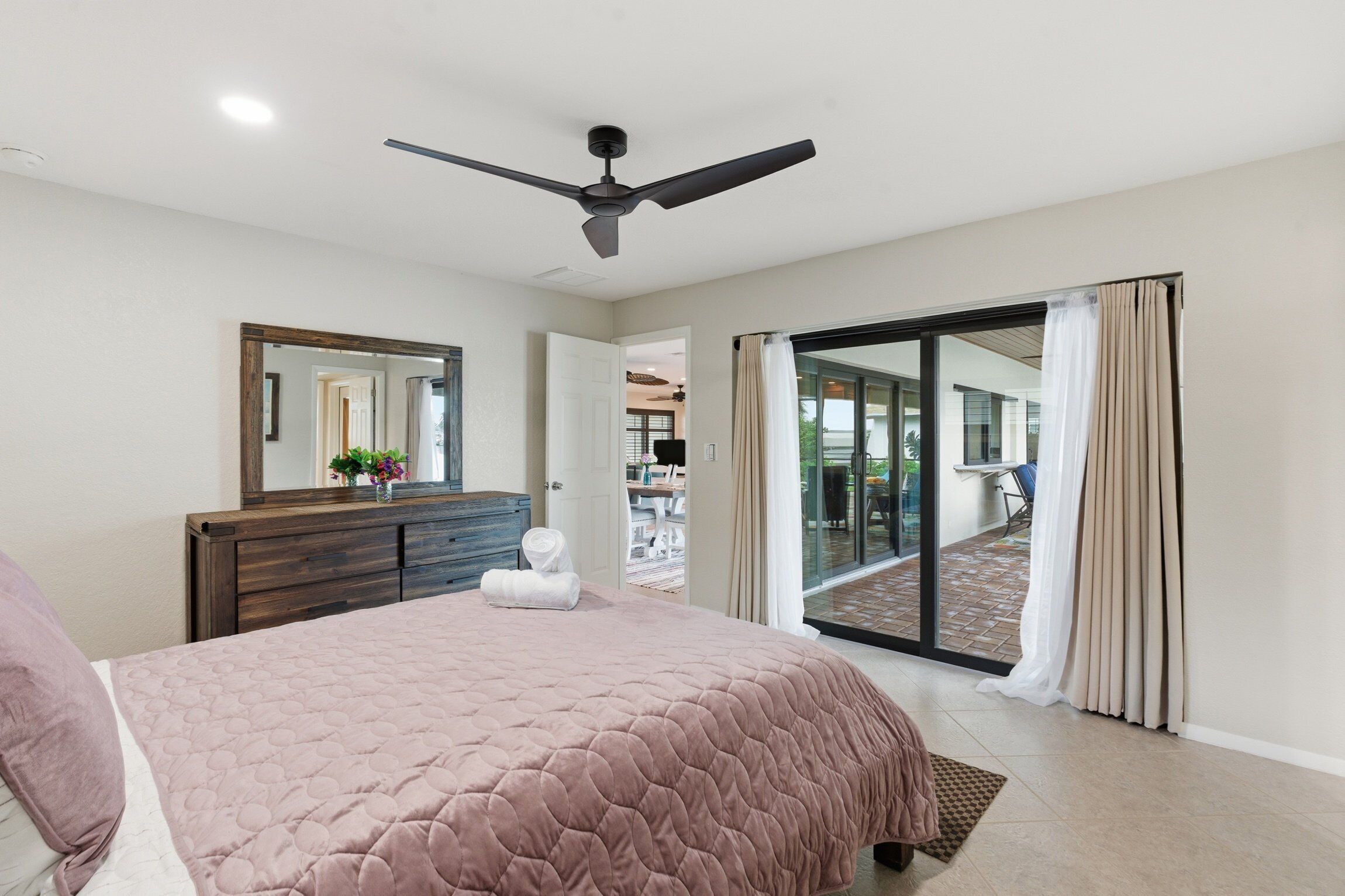 Une chambre à coucher comprenant un lit, un ventilateur de plafond, une commode, un miroir et une porte-fenêtre coulissante donnant sur un espace extérieur.