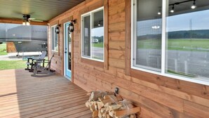 A covered wooden deck with a grill and seating area, large windows, and a view of a road and fields.