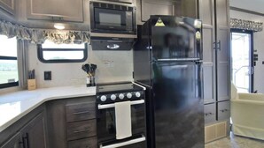 A compact kitchen with a stove, microwave, and refrigerator, featuring wooden cabinets and a window with curtains.