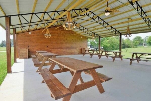 A covered picnic area with wooden benches and picnic tables, under a metal roof with hanging lights.