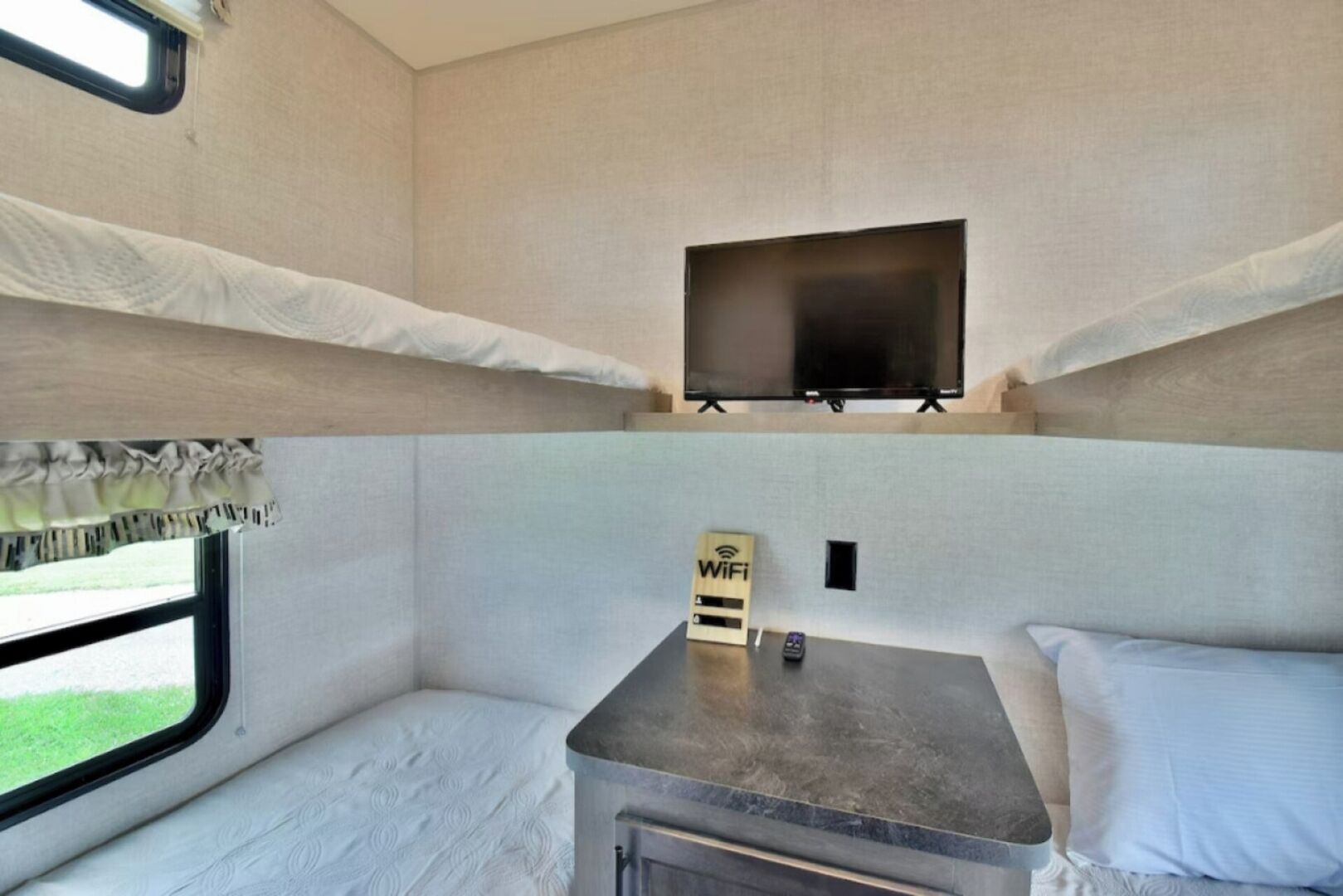 A compact RV interior with a flat-screen TV mounted above a small table, a Wi-Fi sign, and a window with a view of greenery.