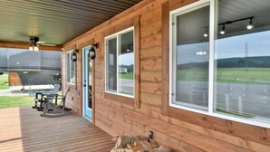 A covered wooden deck with a grill area, large windows, and a view of a grassy field. - Mins from the Lake! Camper King Bed • Firepit (Decatur)