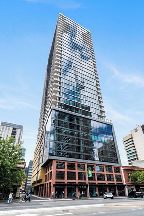 A tall, modern building with a glass facade and a mix of brick and metal elements at the base. - Luxe 1+1 Apt | Huge Balcony+BBQ  | Adelaide CBD (Adelaide)