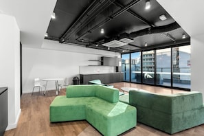 Un espai d'oficina modern amb sofàs verds, una zona de cuina i grans finestres que ofereixen vista a la ciutat.
