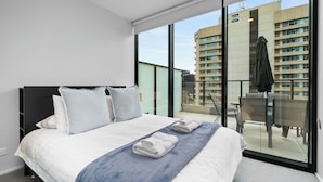 A modern hotel room with a large bed, a balcony with a view of the city, and a dining area with a table and chairs. - Luxe 1+1 Apt | Huge Balcony+BBQ  | Adelaide CBD (Adelaide)