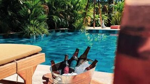Une scène au bord de la piscine, avec un fauteuil de relaxation en bois, un seau rempli de bouteilles de bière et une paire de lunettes posées sur un bloc de bois.