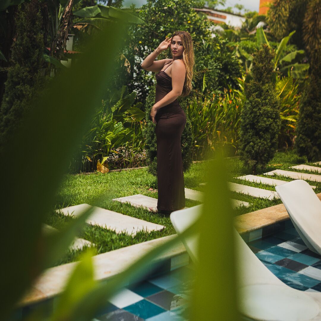 Una persona de pie junto a una piscina, con un jardín y follaje en el fondo.