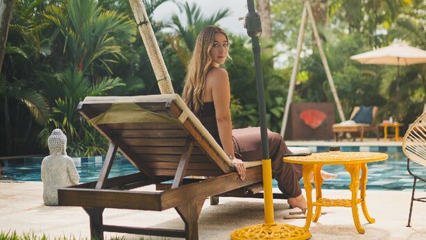 Une personne est assise sur un fauteuil en bois, près d’une piscine, avec une petite table ronde et un parasol à proximité.