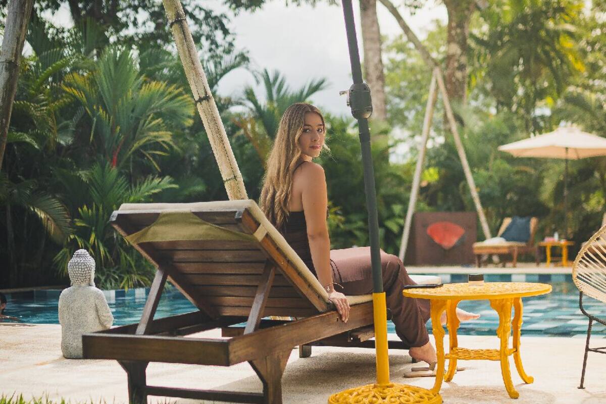 Une personne est assise sur un fauteuil en bois, près d’une piscine, avec une petite table ronde et un parasol à proximité.