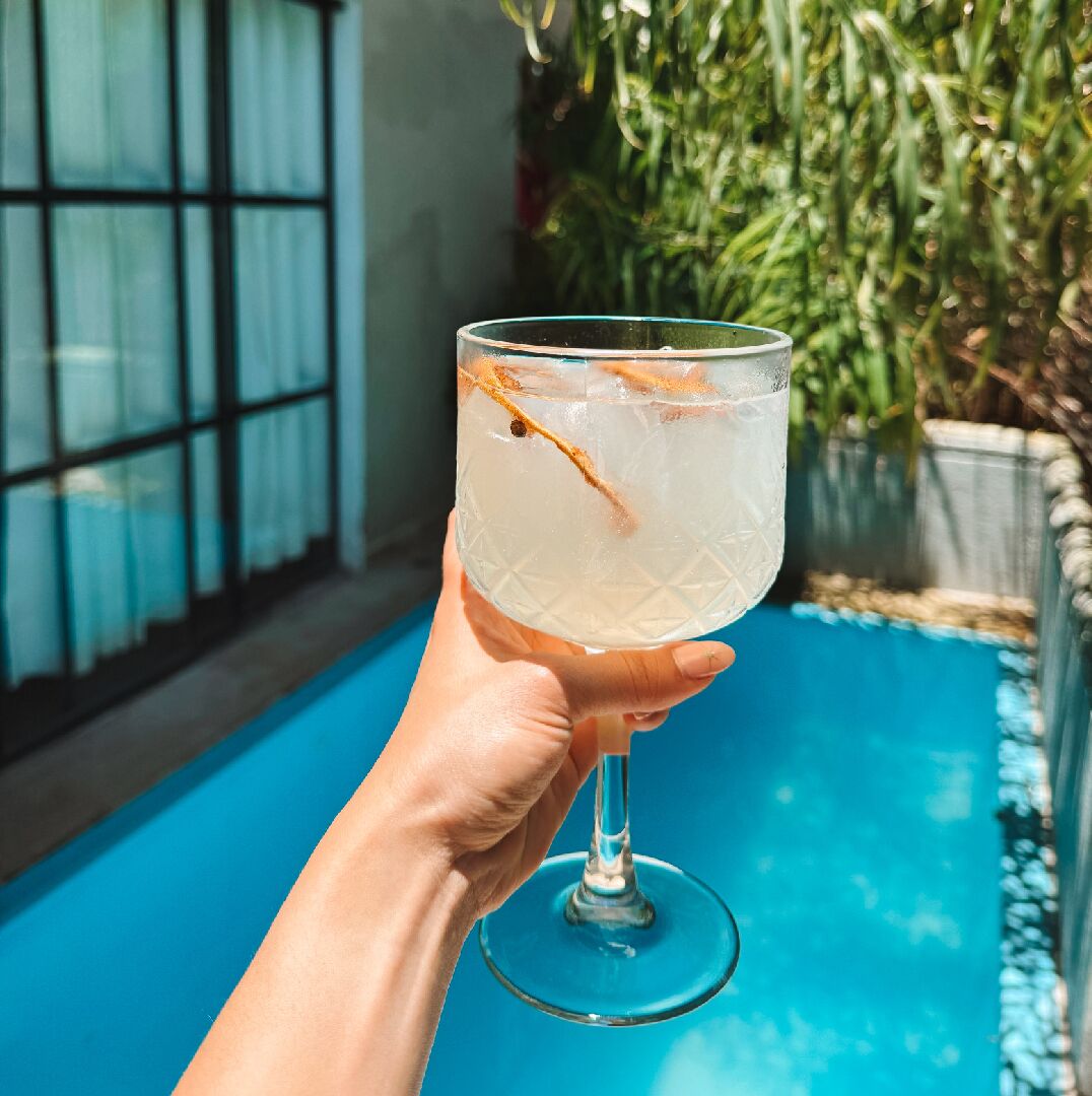 A hand holding a clear glass with ice and a straw, near a pool with a blue floor and a window with blinds.