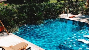 Ein Schwimmbecken mit Schachbrettfliesen, umgeben von üppiger Vegetation und ausgestattet mit Liegestühlen und Sonnenschirmen.