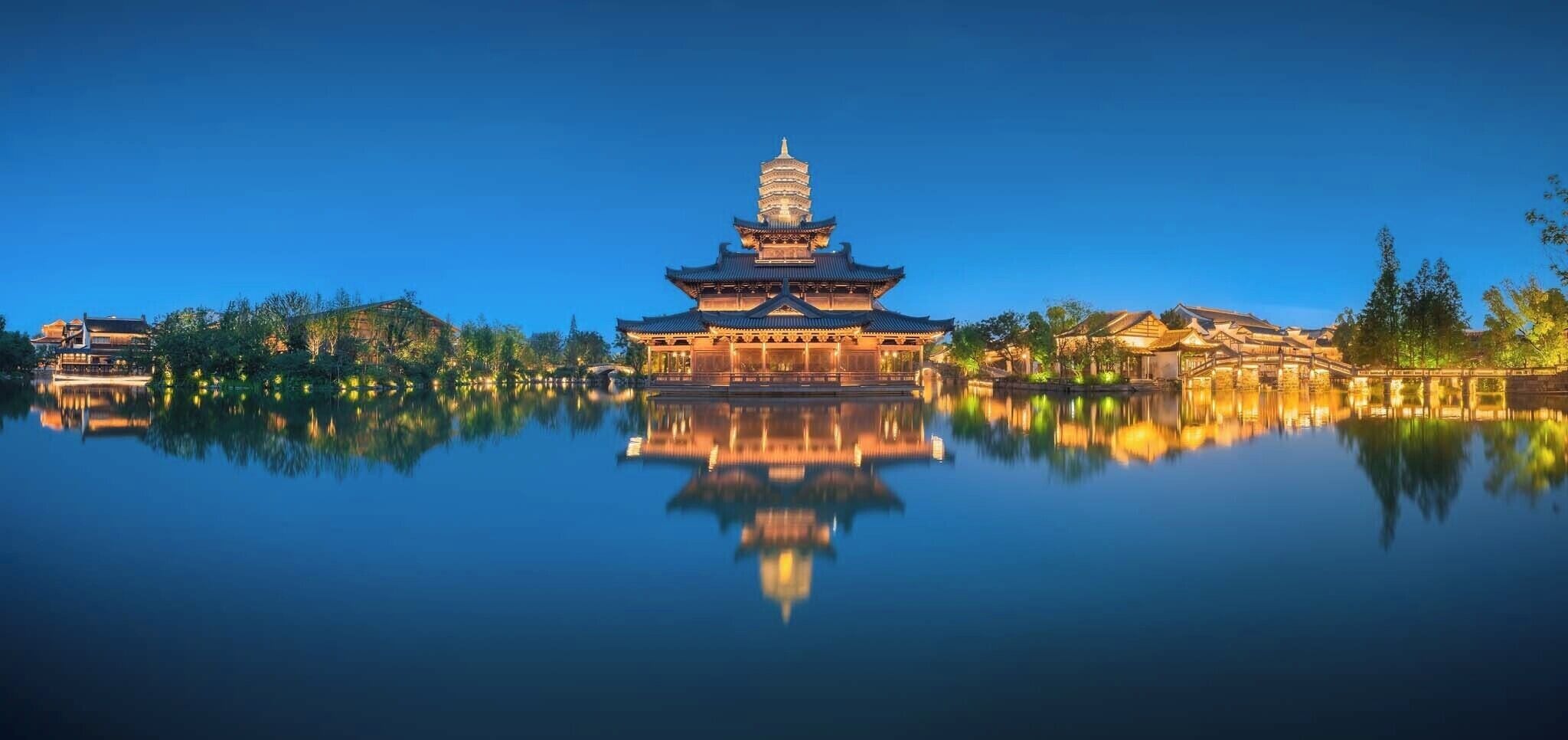 A traditional pagoda-style building with a reflection in a calm body of water, surrounded by trees and other structures.
