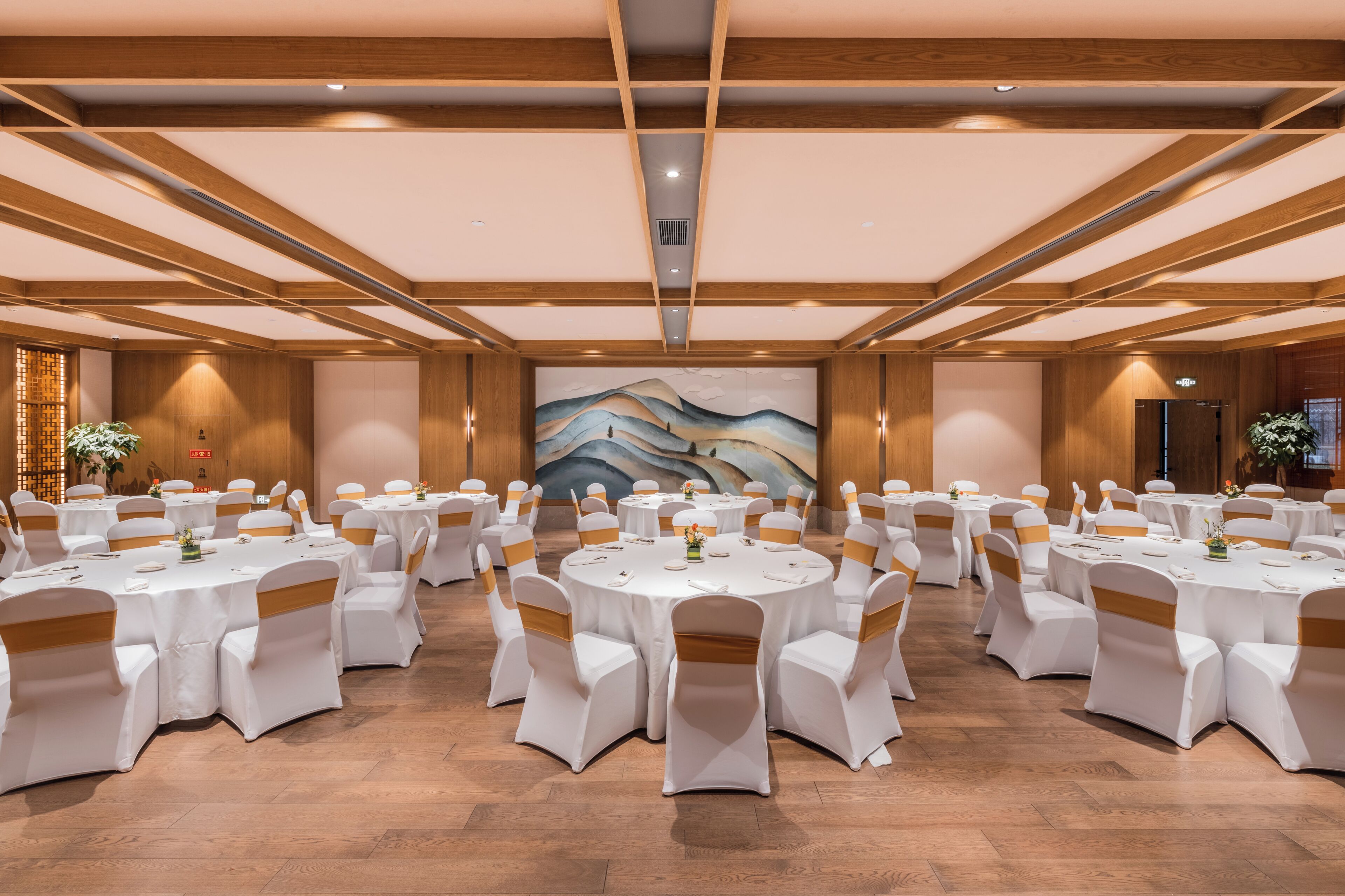 A banquet hall with round tables and chairs, wooden beams on the ceiling, and a large mural on the back wall.