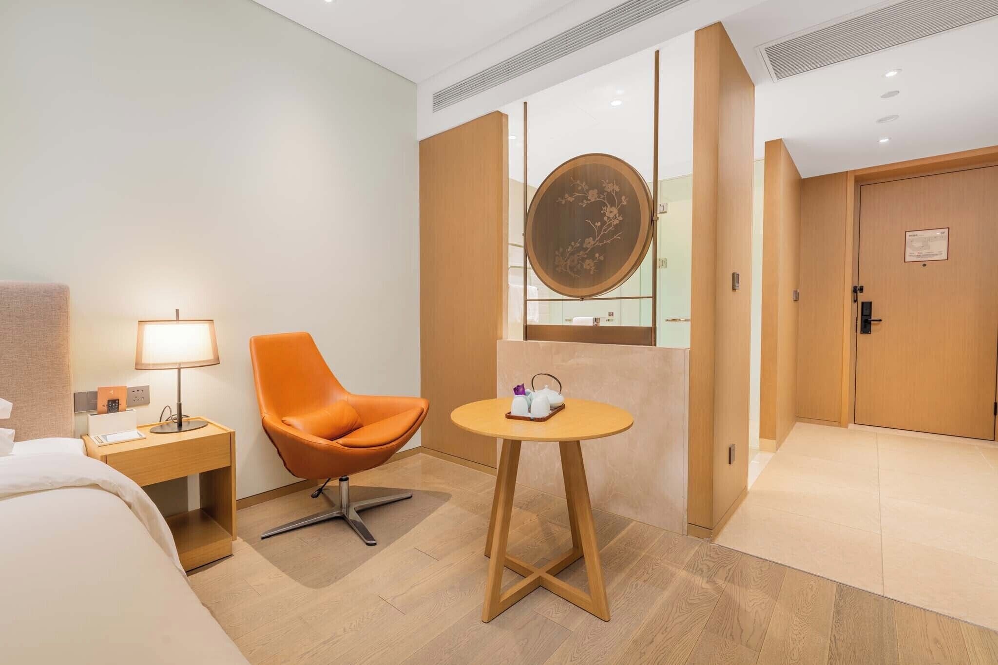 A hotel room with a bed, an orange chair, a small table with a tray, a lamp, and a wooden door.