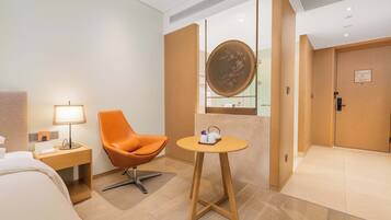 Une chambre d’hôtel avec un lit, un fauteuil orange, une petite table avec un plateau, une lampe et une porte en bois.