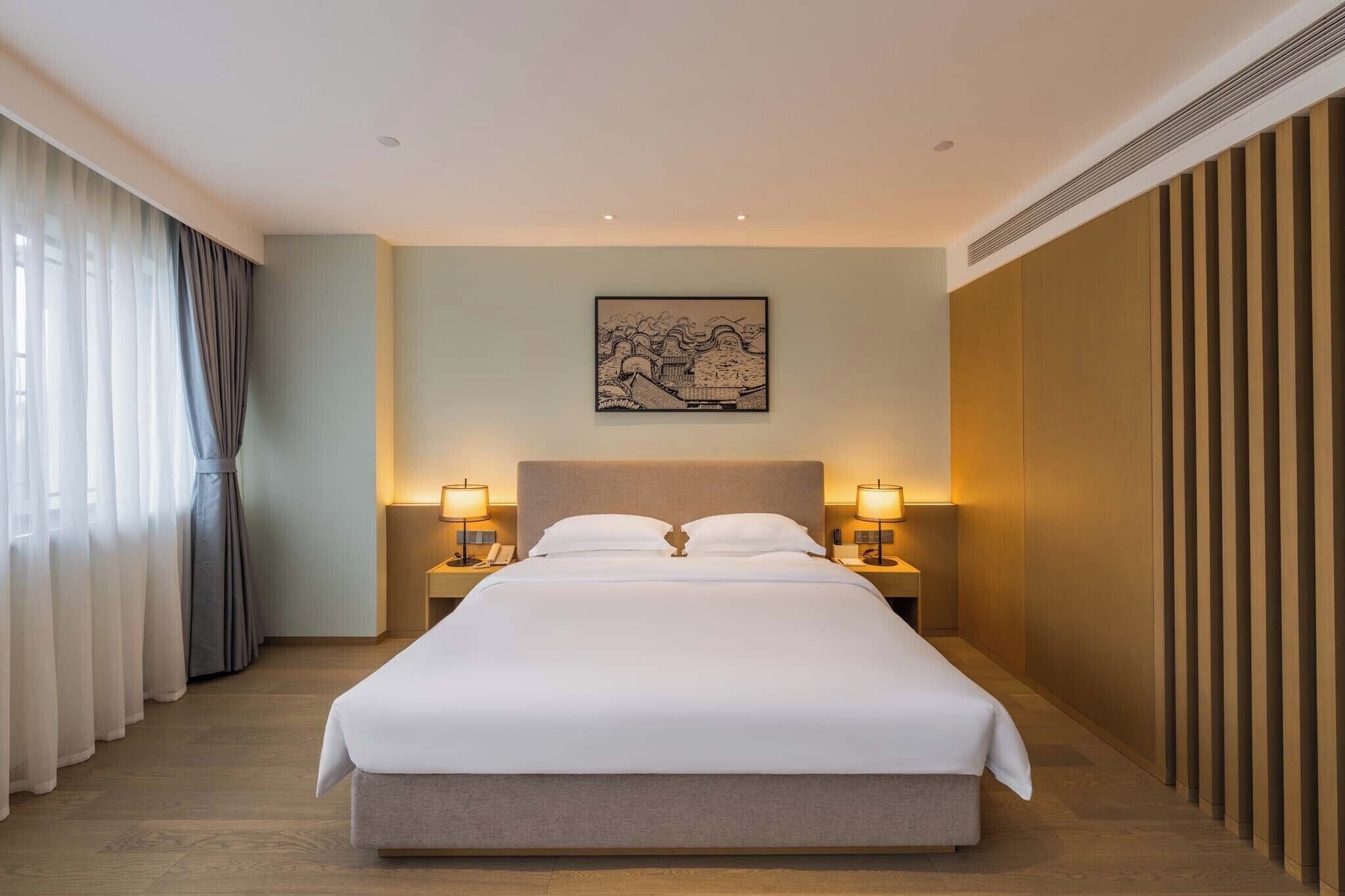 Quarto de hotel com uma cama grande, duas mesas de cabeceira com luminárias, um quadro na parede e vista para o exterior através de cortinas leves.