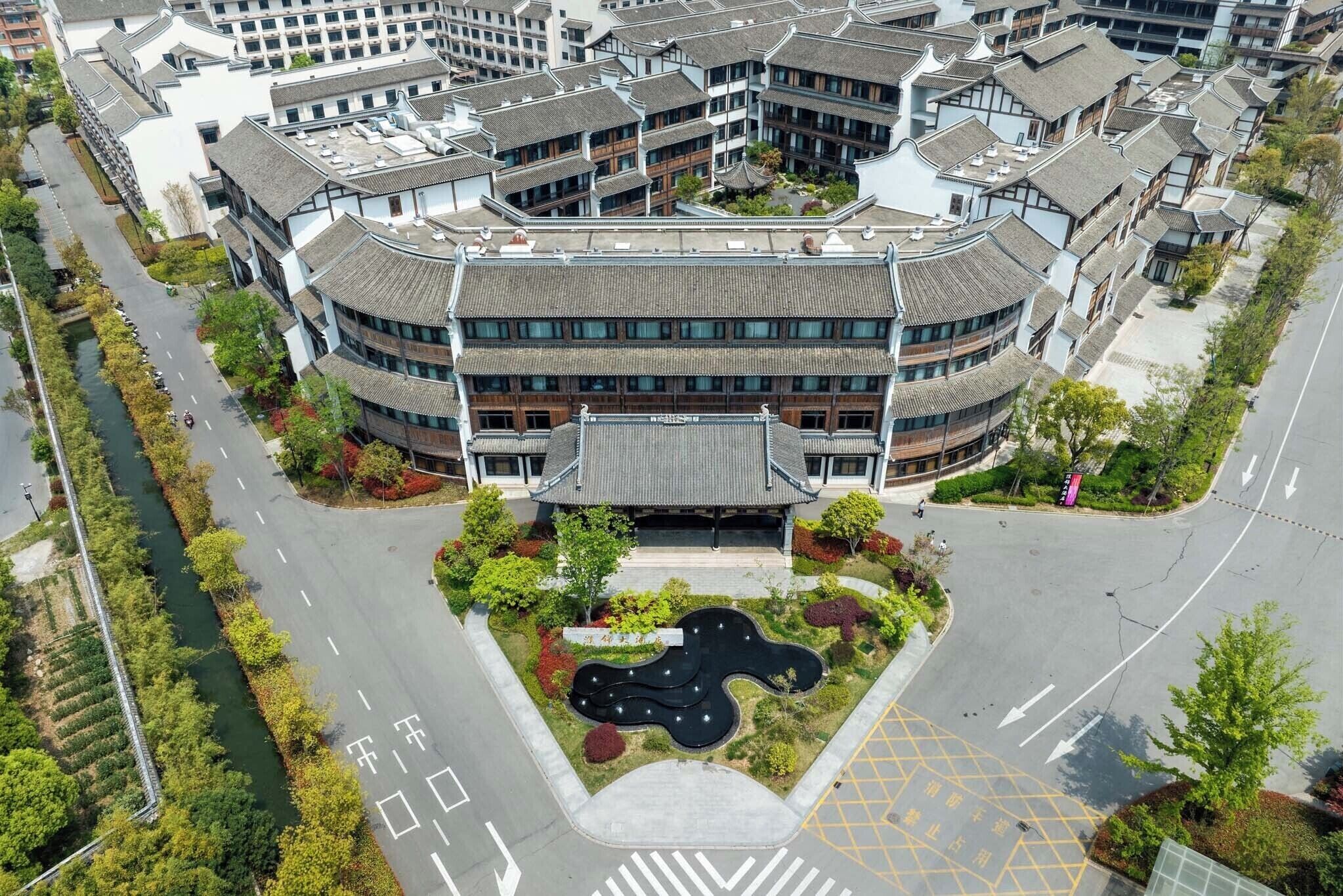 An aerial view of a modern building with traditional architectural elements, surrounded by greenery and a water feature.