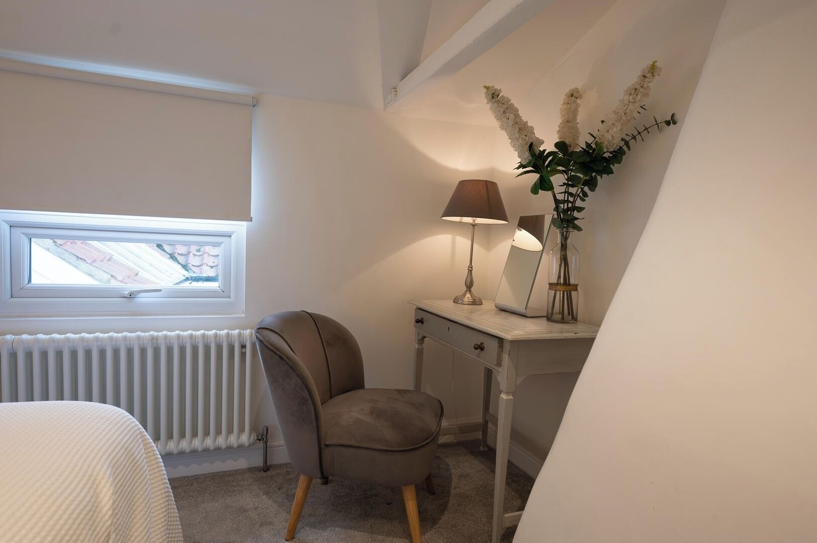 Ett sovrum med ett fönster, en element, en stol, ett skrivbord, en lampa och en vas med blommor.