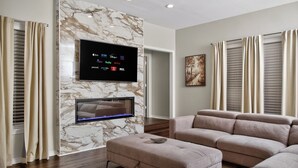 A modern living room with a flat-screen TV mounted on a marble wall, a sectional sofa, and a fireplace. - Luxury Frisco Home w/Private Pool, Game Room, 85" TV, Cozy Patio & Huge Backyard (Frisco)