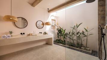 A modern bathroom with a large mirror, a sink with toiletries, a shower, and a plant area.