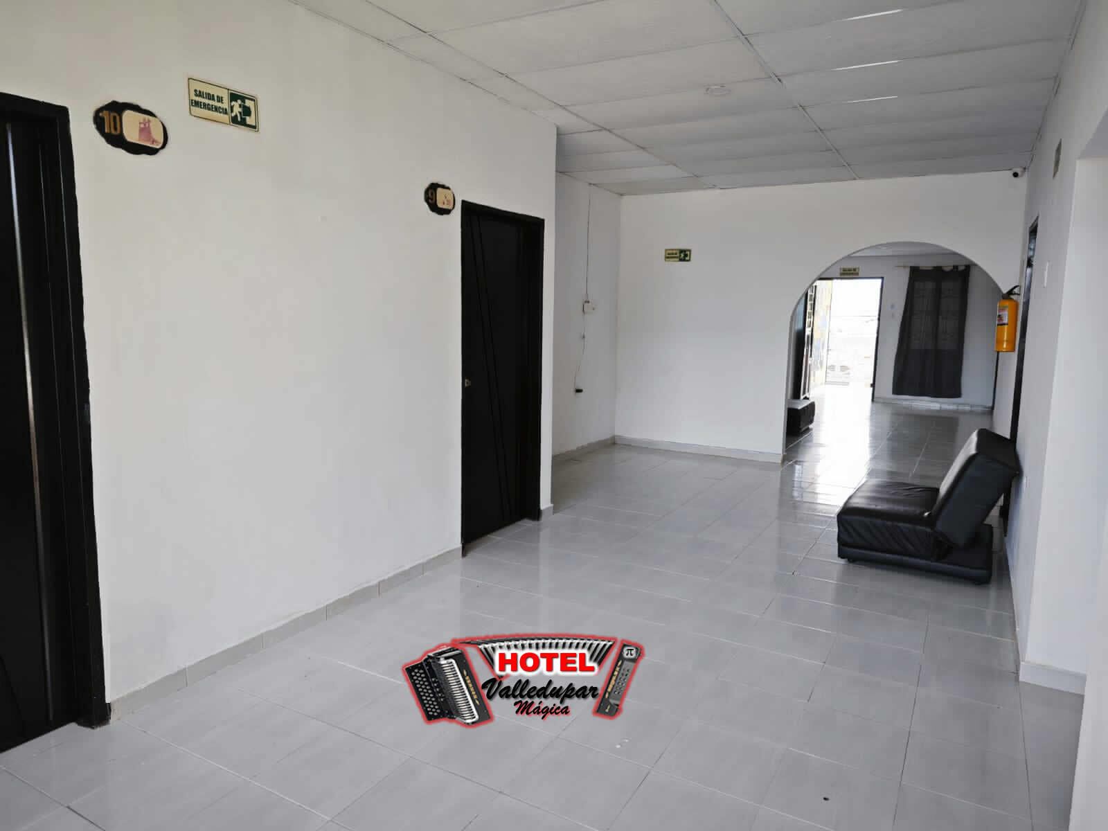 A hotel corridor with a black door on the left, a black sofa on the right, and a sign indicating an emergency exit.