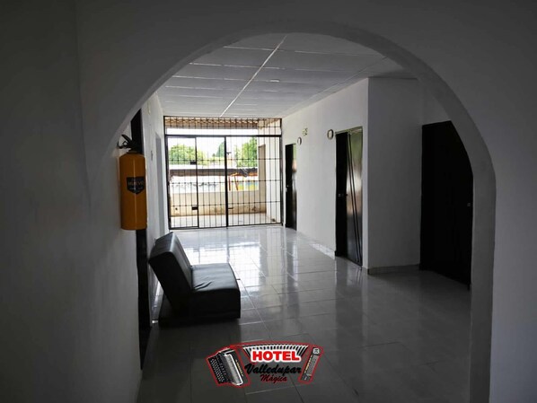 Um corredor de hotel com piso ladrilhado, um extintor de incêndio, um banco e um portão que dá para o exterior.
