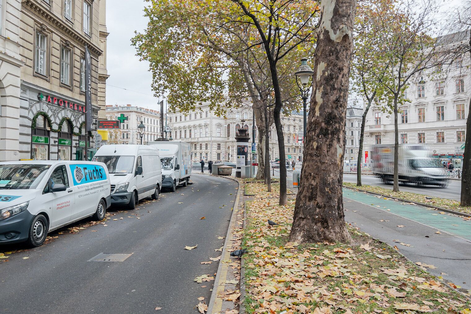 A street with parked delivery vans, trees with autumn leaves, and historic buildings in the background.