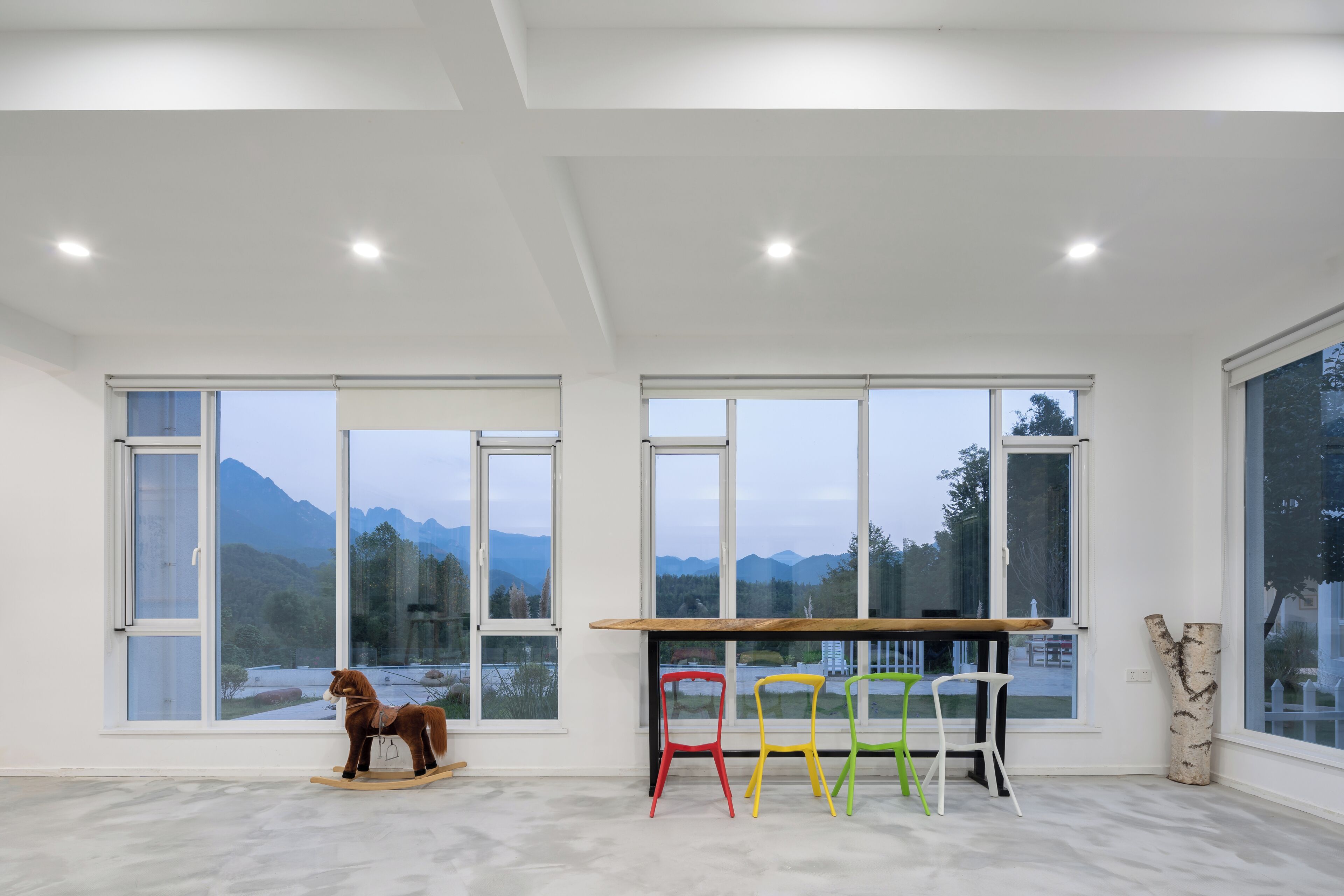 A spacious room with large windows, a wooden rocking horse, and colorful chairs.