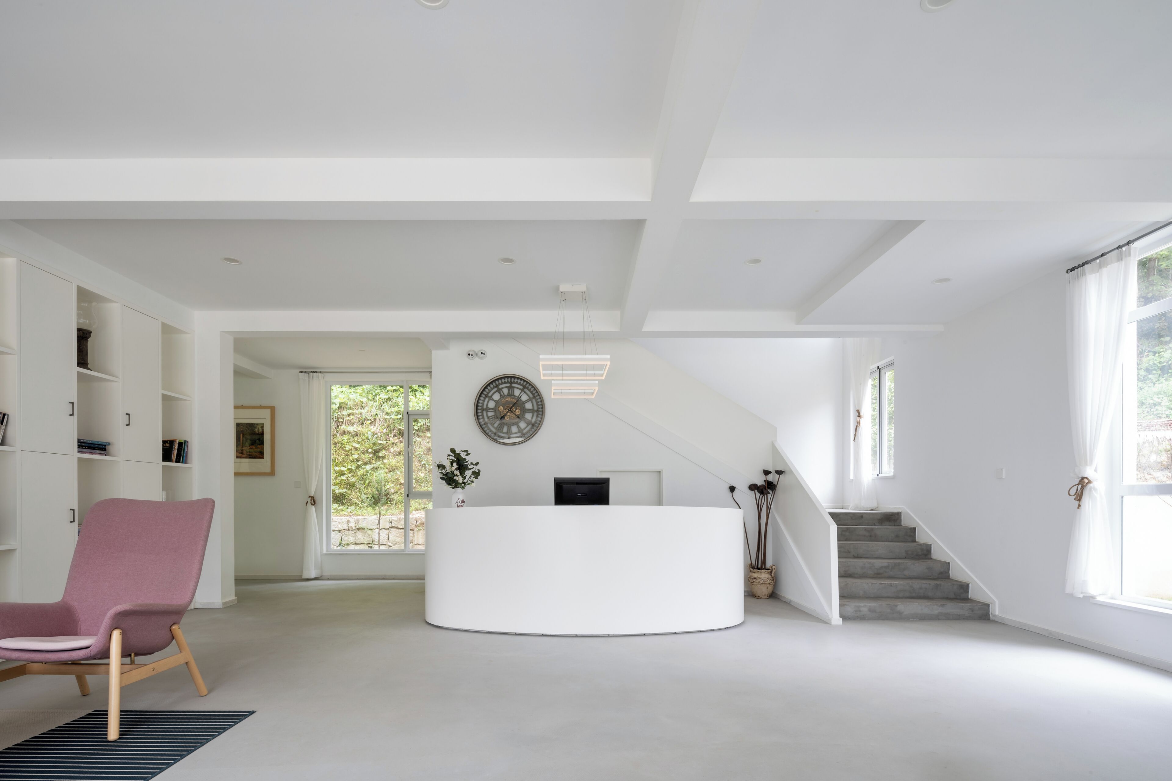 A spacious, modern interior with a central reception desk, a staircase, and a large window.