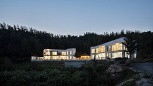 A modern two-story house with large windows and a stone foundation, surrounded by dense forest. - daiyungui (Huangshan)