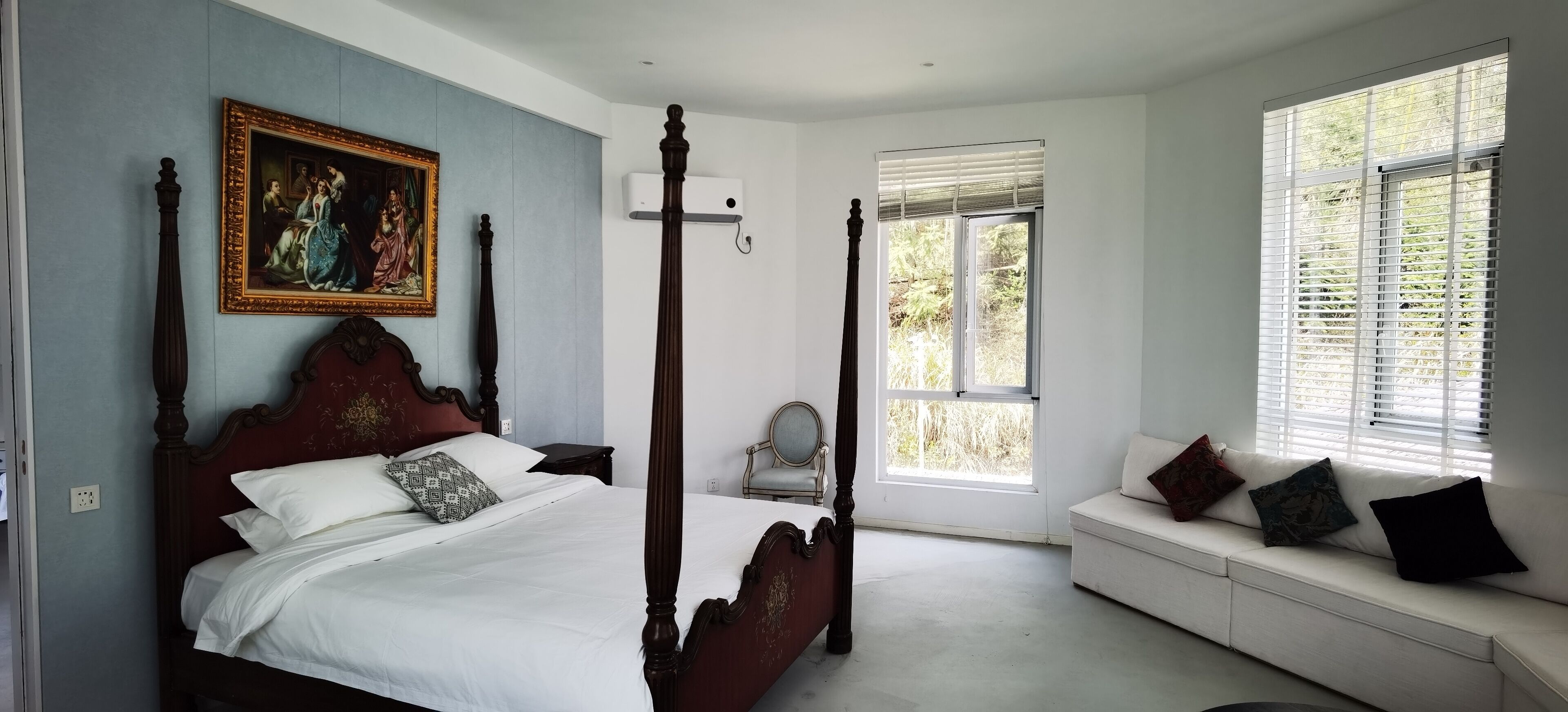 Een slaapkamer met een hemelbed, een groot schilderij, een raam met zonwering, een witte bank met kussens, en een stoel.