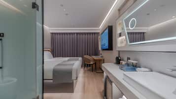 A modern hotel room with a bed, a desk, a TV, and a bathroom with a large mirror and sink.