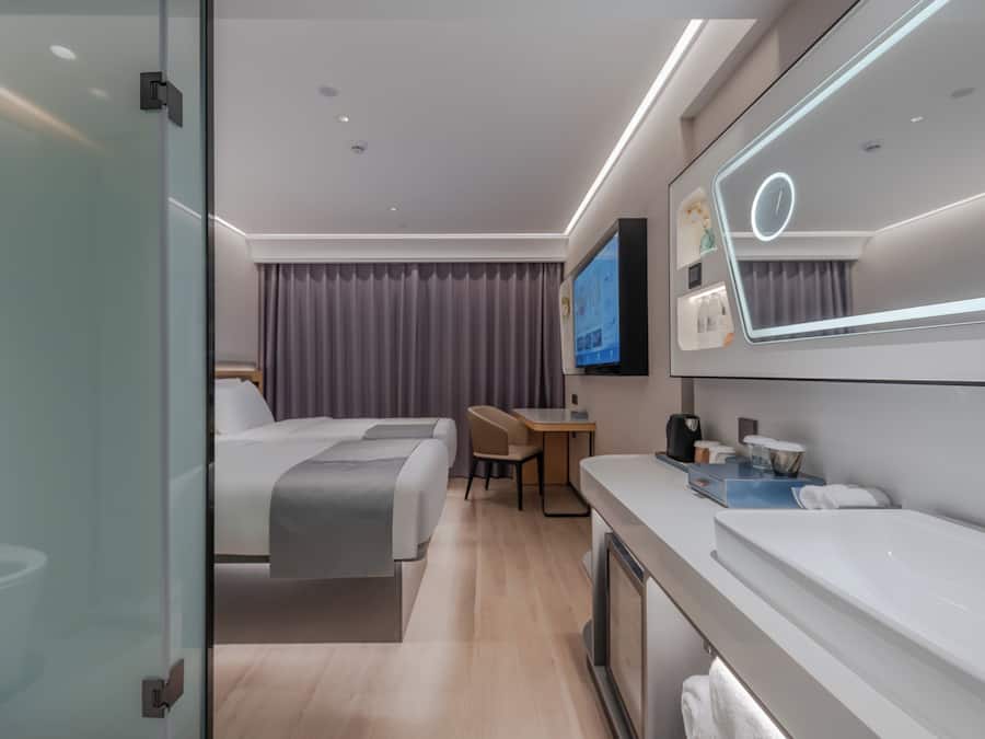A modern hotel room with a bed, a desk, a TV, and a bathroom with a large mirror and sink.
