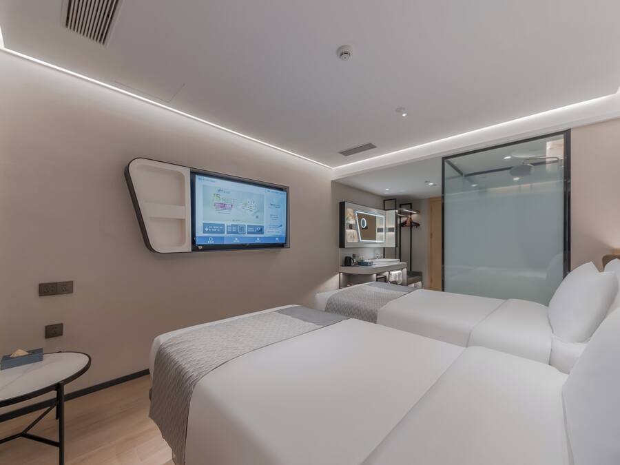 Una habitación de hotel moderna con dos camas, una mesita pequeña, un televisor montado en la pared y un separador de vidrio.