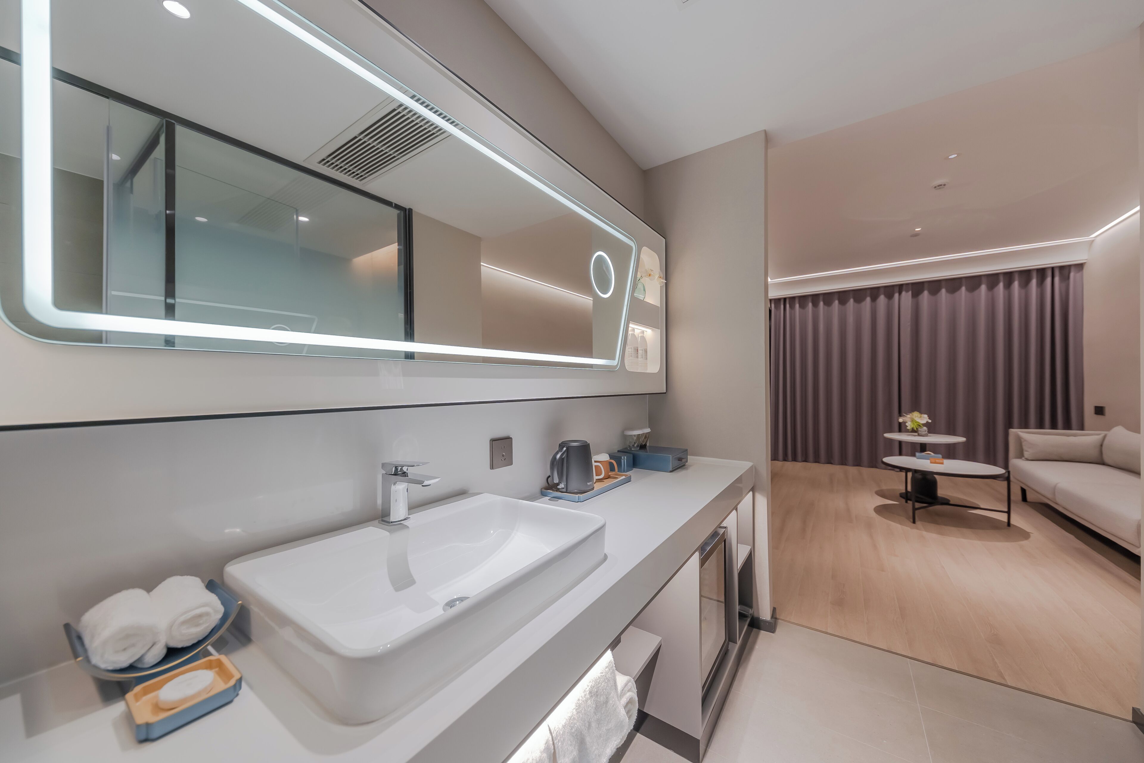 A modern bathroom with a large mirror, white sink, and towel rack. There is a small table with a kettle and a cup.