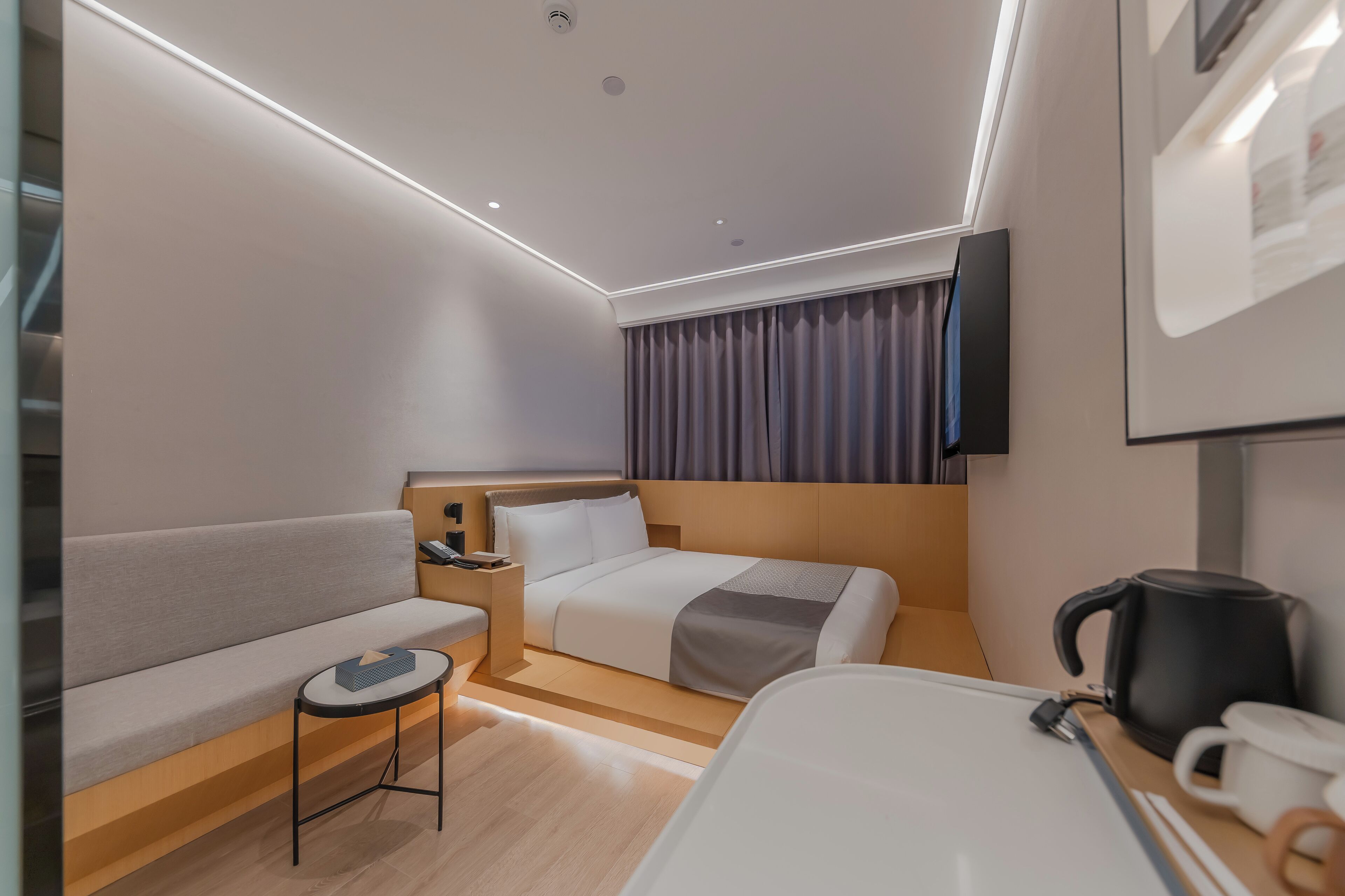 A modern hotel room with a bed, a sofa, a small table, a coffee maker, and a TV on the wall.