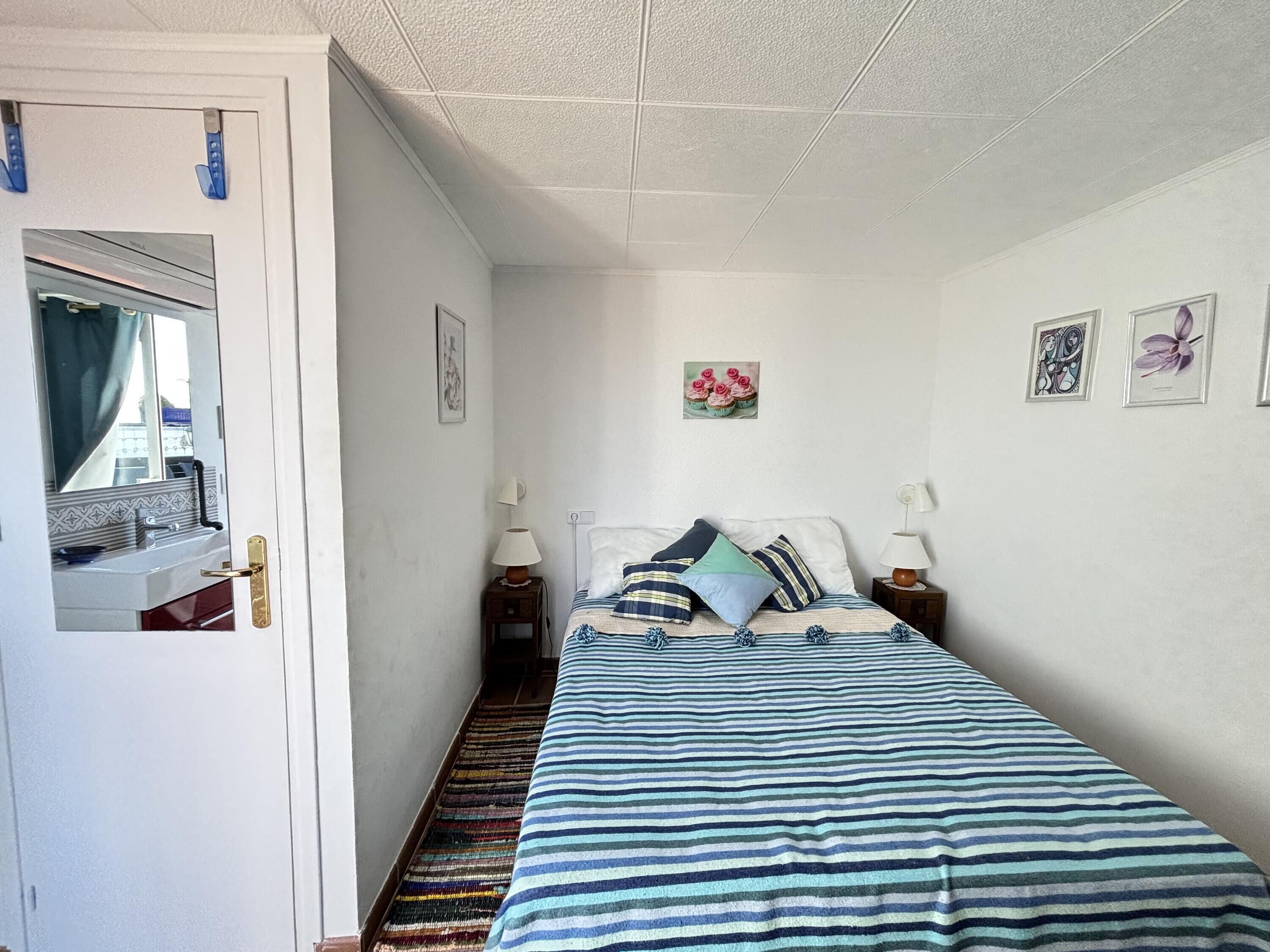 Ein schmales Zimmer mit einem Bett, einem kleinen Tisch mit einer Lampe, einem Spiegel und einer Tür.