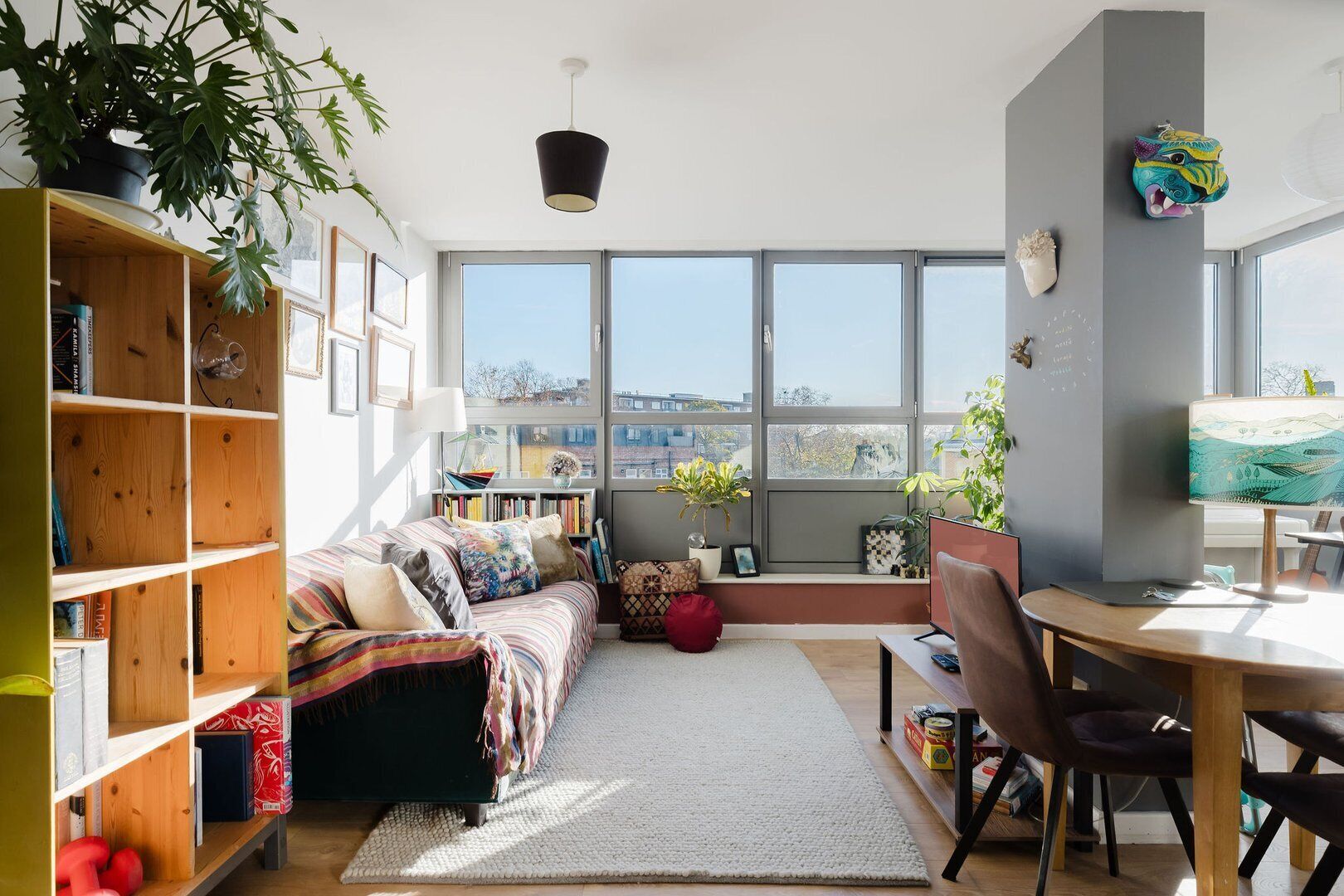 A cozy living room with a sofa, a bookshelf, a dining area, and large windows offering a view of the city.