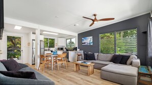A modern living room with a grey sofa, a wooden coffee table, and a grey couch. There is a dining area with a wooden table and chairs, and a kitchen with white cabinets and a microwave.