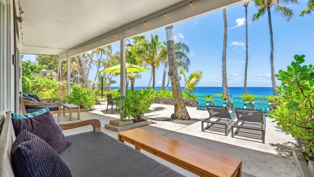 A covered outdoor seating area with a view of the ocean and palm trees.