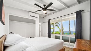 A bedroom with a bed, a ceiling fan, a window with curtains, and a view of the beach.