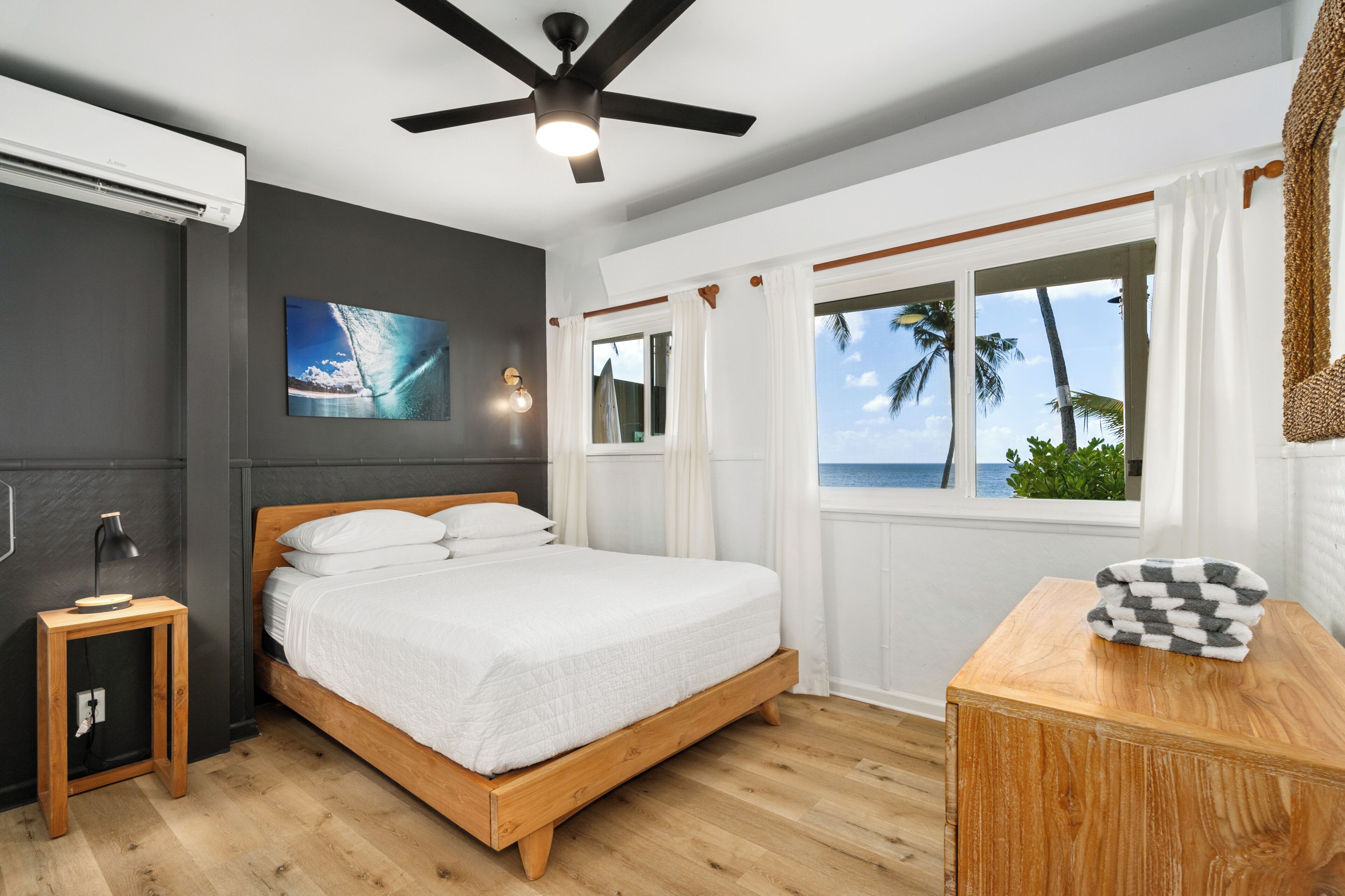 A bedroom with a wooden bed, a ceiling fan, a window with curtains, and a view of the ocean.