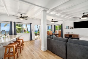 A modern living space with a kitchen area, wooden bar stools, a gray sofa, ceiling fans, and a view of the ocean.
