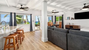 Ein modernes Wohnzimmer mit Küchenbereich, Holzbarhocker, einem grauen Sofa, Deckenventilatoren und Meerblick.